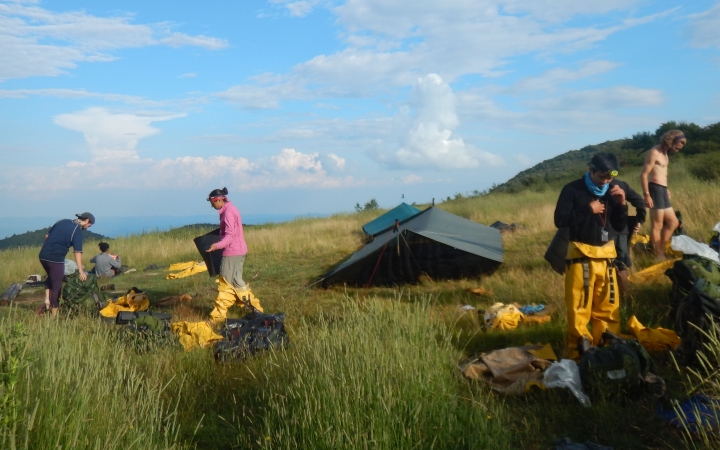 A couple of tarp shelters rest in a grassy meadow under blue skies. A few students stand nearby the shelters. 